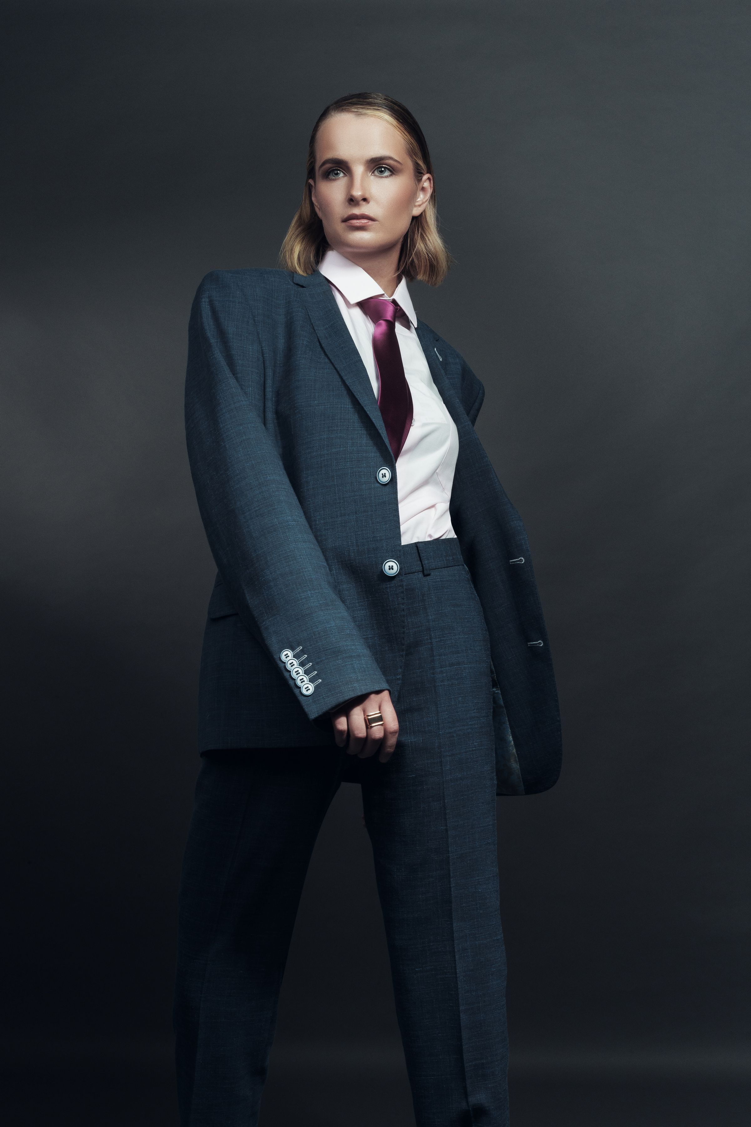 Editorial fashion portrait of a woman in a tailored blue suit and burgundy tie against a charcoal studio backdrop
